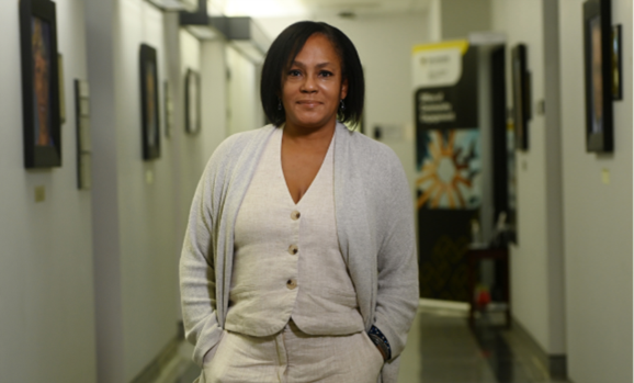 A woman stands in a hallway wearing a white vest, pants and grey sweater, smiling at the camera.