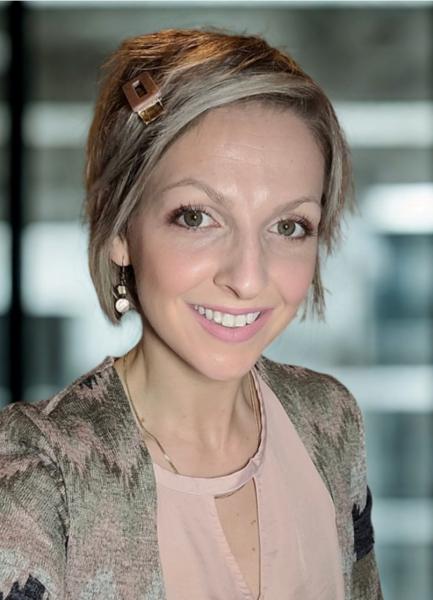 A woman with short, blonde hair wears a pink shirt with a printed sweater over top and is smiling at the camera.