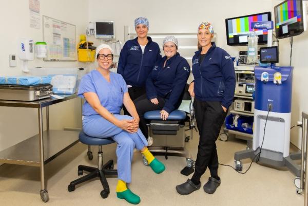 A group of four medical professionals sit in a hospital room wearing scrubs and surrounded by medical equipment.