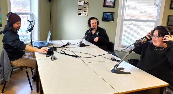 Three people wearing headsets are seated indoors in a room at a table with microphones. 