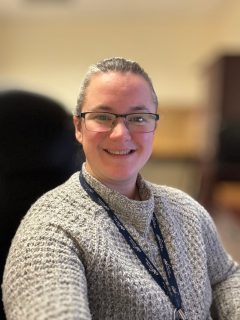 A woman with a grey sweater. Her hair is pulled back and she is wearing a landyard around her neck
