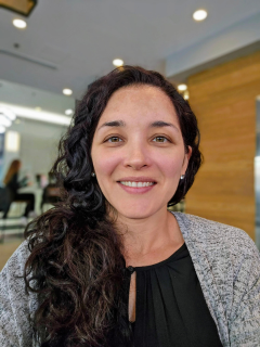 A woman with long brown curly hair. She is wearing a grey cardigan over a black blouse.