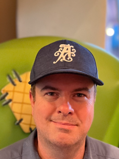 A man with a blue baseball cap on. He is wearing a dark grey collared shirt and has black sunglasses hanging on his collar.
