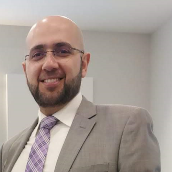  A man wearing a light gray suit jacket, a white dress shirt, and a purple patterned tie stands indoors against a plain, light‑colored wall with minimal décor.