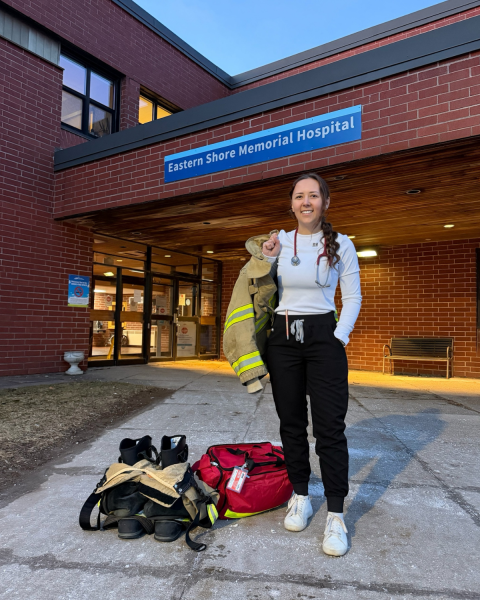 Image of a nurse in black and white scrubs, wearing a stethoscope around her neck and a firefighter jacket over her right shoulder. She is facing the camera smiling. She has fire fighter pants and boots, plus a medical bag next to her feet. She is standing outside a hospital "Eastern Shore Memorial Hospital."