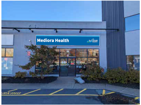 Photo taken outside of a Nova Scotia Health clinic. Plain grey front with blue sign that says "Mediora Health." Taken from a parking lot.