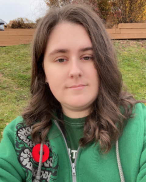 A photo of a woman outside with brown wavy hair. She is wearing a green zip-up sweater with a grey design on the left shoulder and a red poppy pin.