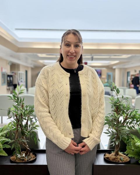 A woman stands in a room wearing a white cardigan, black shirt and grey pants, smiling at the camera.
