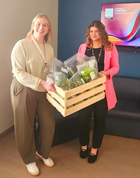 Two female healthcare workers are featured in this photo. They are smiling and holding a crate of fresh greens. 
