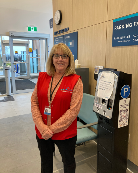 A woman stands in the lobby of a healthcare facility wearing a red vest, pink shirt, black pants, glasses and a smile.