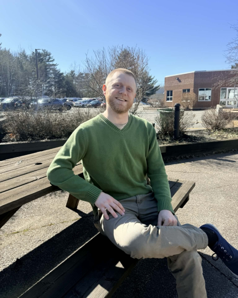 A man sits outside on a picnic table wearing a green shirt and beige pants, smiling at the camera.