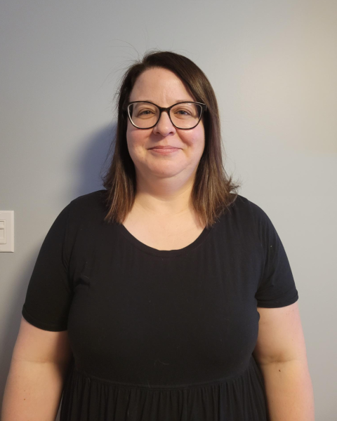 A woman with long, brown hair stands against a grey wall wearing a black shirt and glasses.