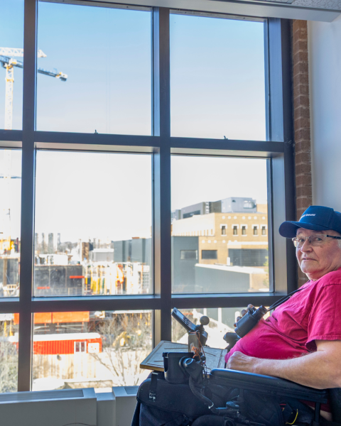 A man sits in a wheelchair with a window behind him with a view of a construction site and a crane.