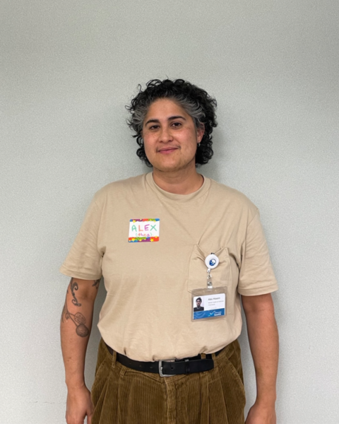 A person with short, curly hair, wearing a beige shirt and brown pants stands in front of a white wall, smiling at the camera.