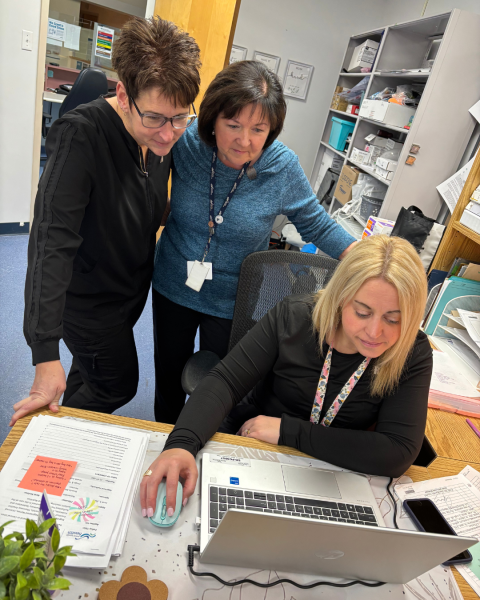 3 healthcare workers, 2 standing and 1 sitting in front of a laptop in an office.