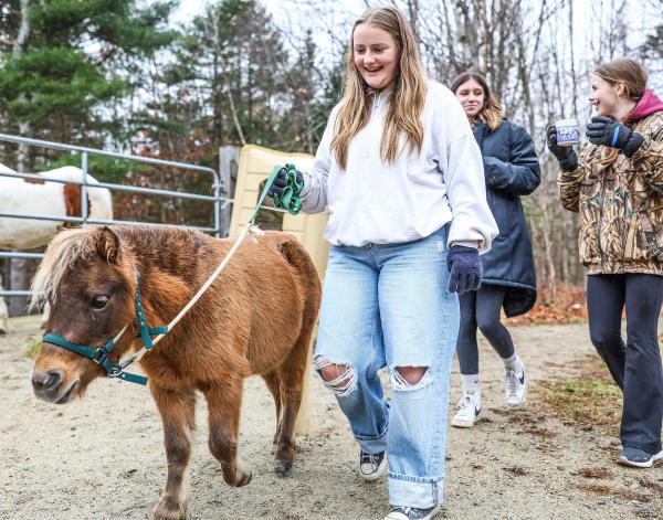 A smiling young woman walks a small pony. Two other young women are in the background. The scene is set outdoors with trees visible in the background. 