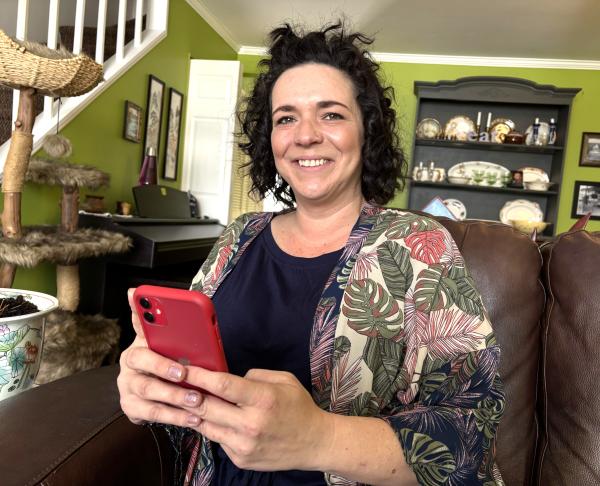 Woman sitting on a couch at home holding a smartphone and smiling, with a cat tree and home décor in the background, suggesting she is comfortably checking information on a mobile app.