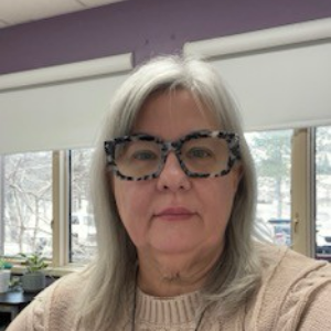 A woman with grey long hair. She is wearing black framed glasses with a brown sweater.