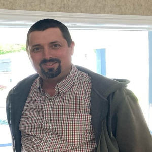 A man wearing a grey hoodie over top a plaid button up shirt and brown khakis. He has short brown hair and a brown goatee.