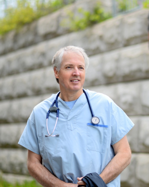 Picture of a male physician in light blue scrubs with stethoscope around his neck. He has grey short hair and is smiling. The picture is taken outside with a rock wall scenery outdoors. 