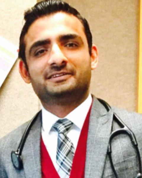 Photo of a man with short dark hair, smiling. He is wearing a grey sports jacket and a red vest underneath with a white collared shirt and a black and white striped tie.