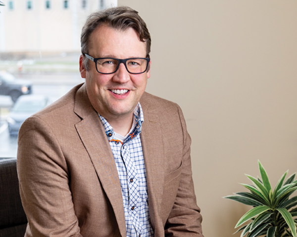 Image of a man with light brown hair and glasses. He is wearing a brown sports coat and blue and white checkered shirt. He is smiling largely and he is seated in an office.