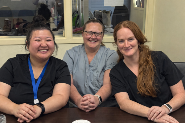 Three women sit together on a couch wearing t-shirts, smiling at the camera.