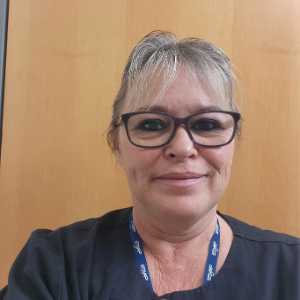 A woman with light hair tied back. She is wearing black framed glasses and a navy blue buttoned blouse. She has a blue NSH lanyard around her neck.