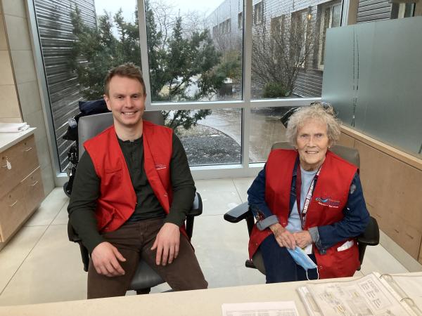 A man and woman sit side-by-side in chairs at a healthcare facility wearing red vests and smiling at the camera.