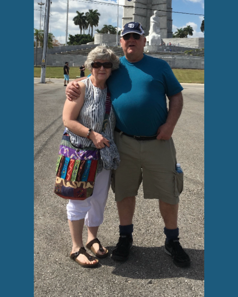 Picture of a (R) man and woman (L) both wearing sunglasses with a picturesque background outside of a statue and palm trees. 