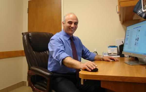 A man sits in a chair wearing a blue-collared shirt and a red tie in front of a computer screen and is smiling at the camera.