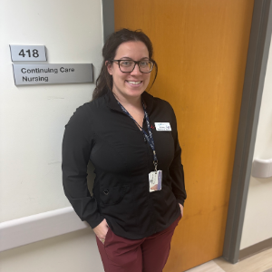 A woman with brown hair tied in a ponytail. She is wearing black framed glasses with a black sweater and maroon pants. She has a NSH lanyard around her neck and a name tag clipped on her sweater.