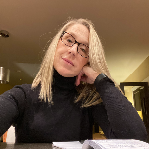 A photo of a woman with straight blonde hair wearing black framed glasses and a long sleeve black turtle neck
