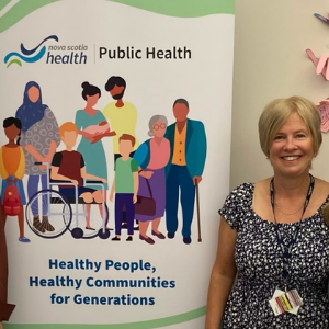  A woman with short blond hair. She is wearing a black NSH lanyard with a flowered black and white top. She is standing in front of a Public Health sign.