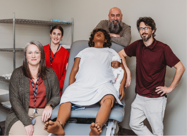 2 female healthcare providers on left side of image, there is a patient mannequin in the middle with a hospital gown on sitting on a chair. There is a male healthcare worker behind the mannequin and another male healthcare worker to right side of mannequin.