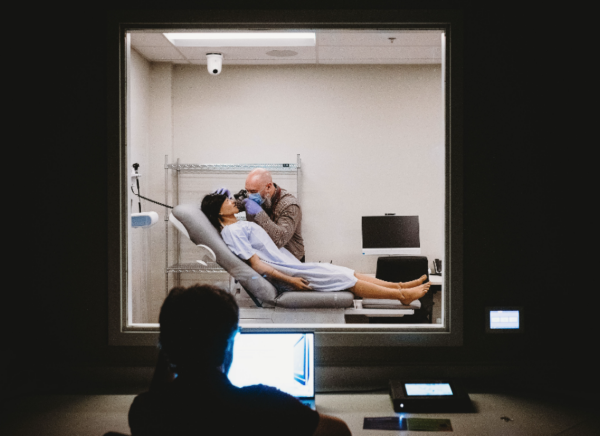 Image is taken looking through a window. A person on one side of the window is watching and has a computer screen in front of them. The person behind the other side of the window is a healthcare worker doing an exam on a mannequin. 