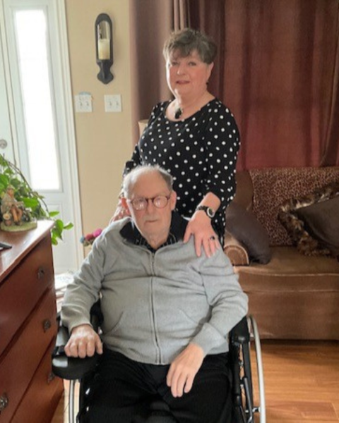 Older couple in their living room. The woman is standing behind her husband with her hand on his shoulder. The man is sitting in a wheelchair.