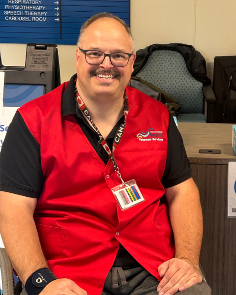 A bald man in a red vest, black short-sleeve shirt sits in a healthcare facility smiling at the camera.