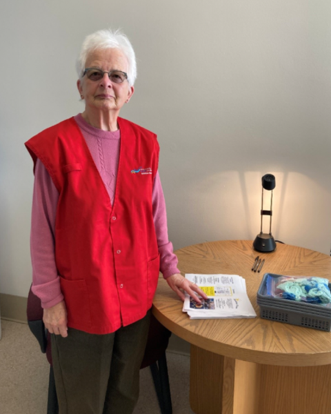 A woman with short, white hair wears a red vest and pink long-sleeve shirt stands in a hospital room beside a table, smiling at the camera.