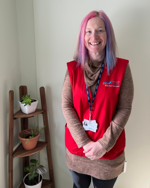A woman with pink and blue hair stands in a room wearing a red vest next to shelf with plants on it, smiling at the camera.