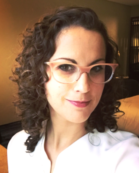 A woman with long, brown, curly hair wears glasses and a white shirt, smiling at the camera.