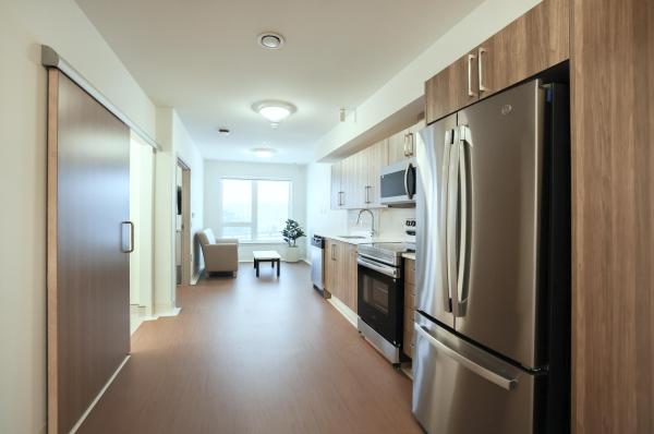 Image looking into a room with wide hallway, it is a view of an apartment style room. You see the kitchen with stainless steel appliances and a view into the living room.