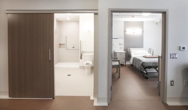View looking into two separate rooms, one is a hospital style room with a bed and bedside table, the other on the left side is a bathroom view.
