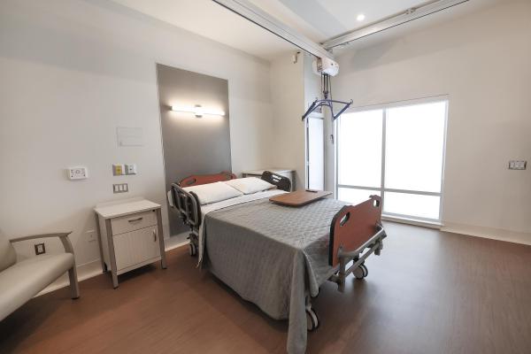 There is a large hospital bed for bariatric patients with a ceiling lift above the bed. There are bright windows and a bedside table and oversized armed chair.