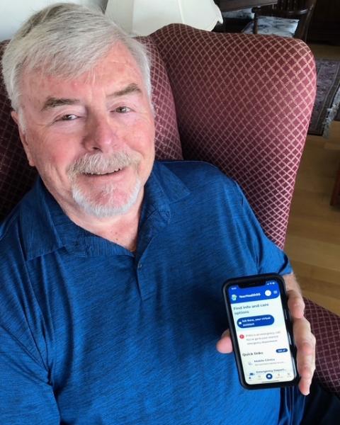 An older man with gray hair and a trimmed beard sits in a red patterned armchair, smiling at the camera while holding a smartphone that displays a healthcare app with options for finding care and services.