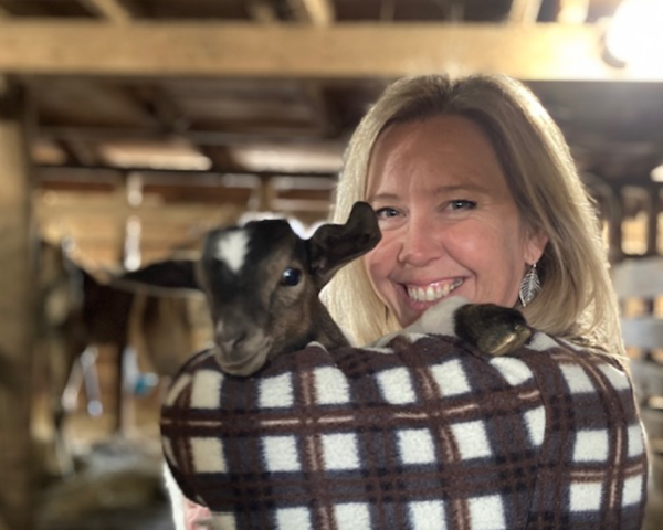 Photo of a woman with blonde woman wearing a brown and white checkered jacket holding a baby goat in her arms.
