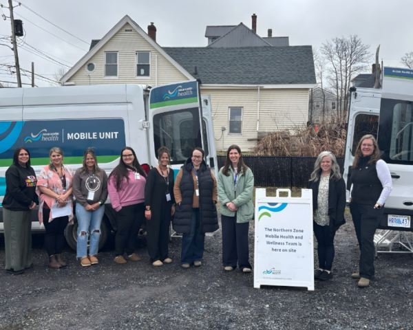 Image of 9 women standing in front of a van that says Nova Scotia Health mobile unit outside. There is a sign propped in between the 7th and 8th woman that says "The Northern Zone Mobile Health and Wellness Team is here on site."
