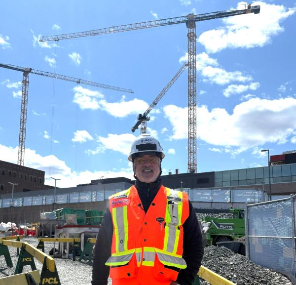 A man stands in front of a construction site wearing an orange safety vest and hard hat.
