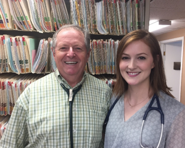 Photo of a man (L) and woman (R) standing in a room with medical charts/folders behind them. They both are smiling standing next to each other very professional with stethoscope's around their necks.