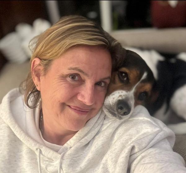 A woman with her dog in a home. She has blonde hair and is wearing a white hoodie. Her dog is black, brown and white and their head is on her shoulder. 